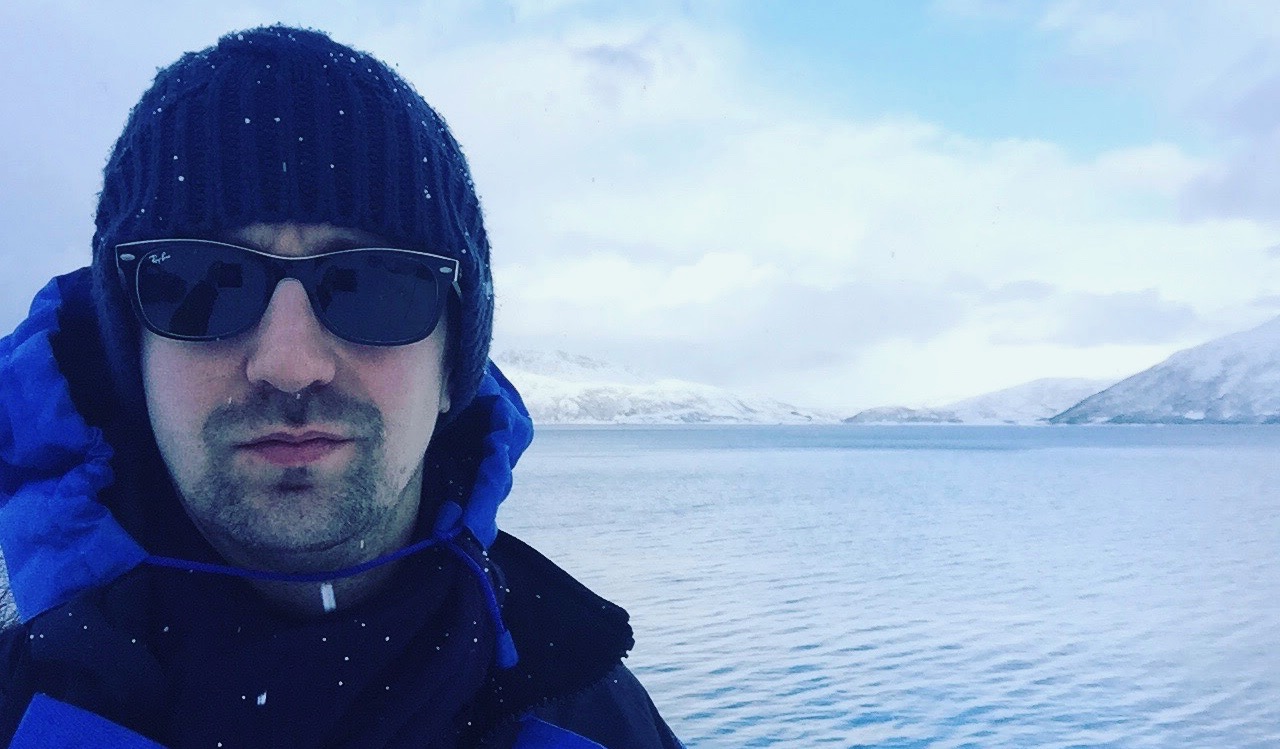 David standing in front of the water with snowy mountains behind him